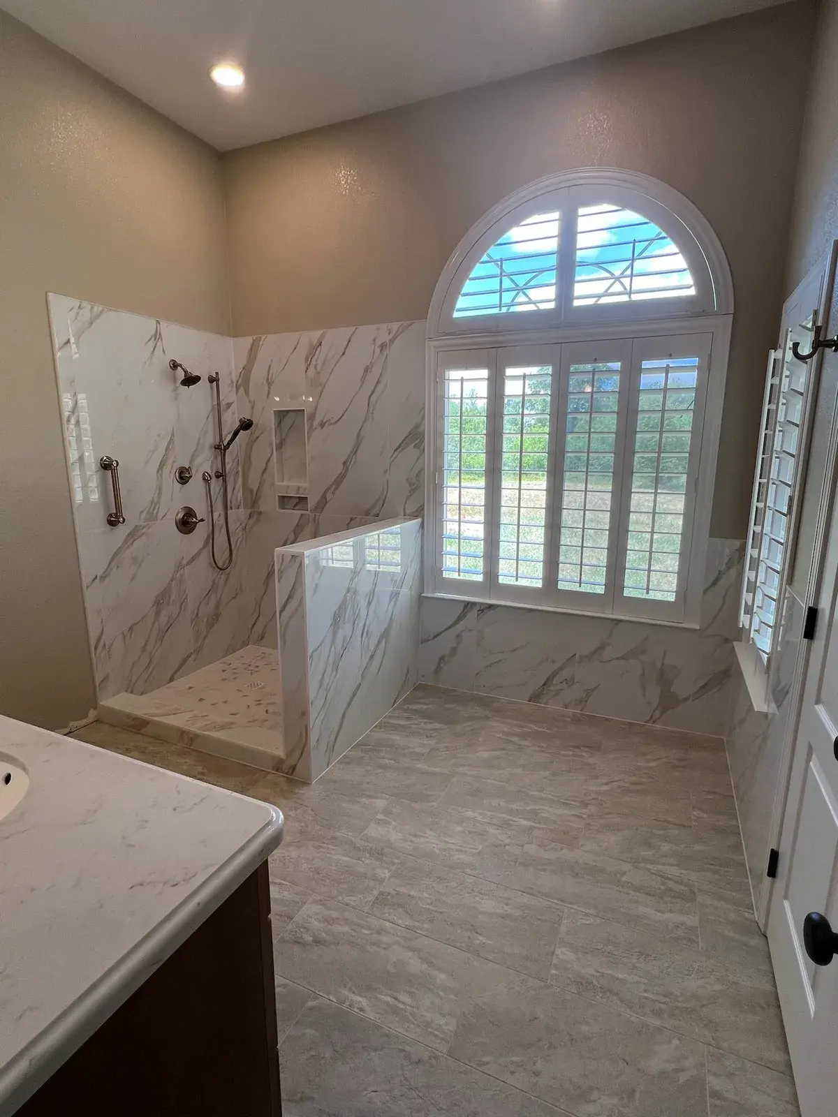 Marble doorless walk-in shower beside an arched shuttered window and vanity