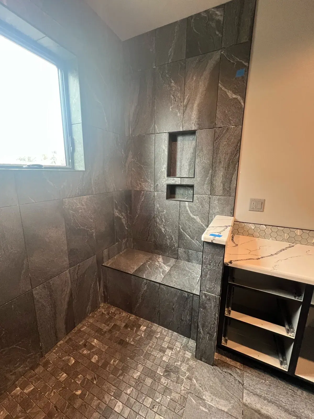 Master bathroom shower in dark gray marble-look tile with a pebble-mosaic floor and quartz vanity